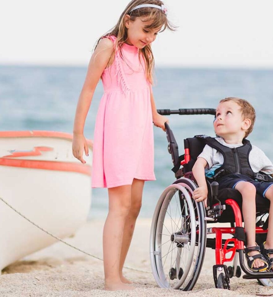 A girl standing beside a young boy using a wheelchair at the beach